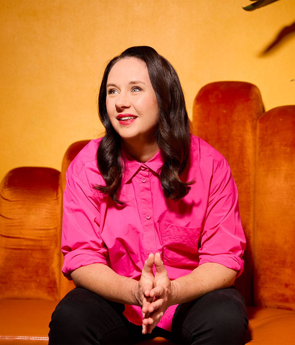 Comedian Mel Buttle sits on an orange velvet sofa against a warm golden-orange background, wearing a bright pink button-up shirt and black trousers. She smiles while looking to the side, her hands clasped together in front of her.